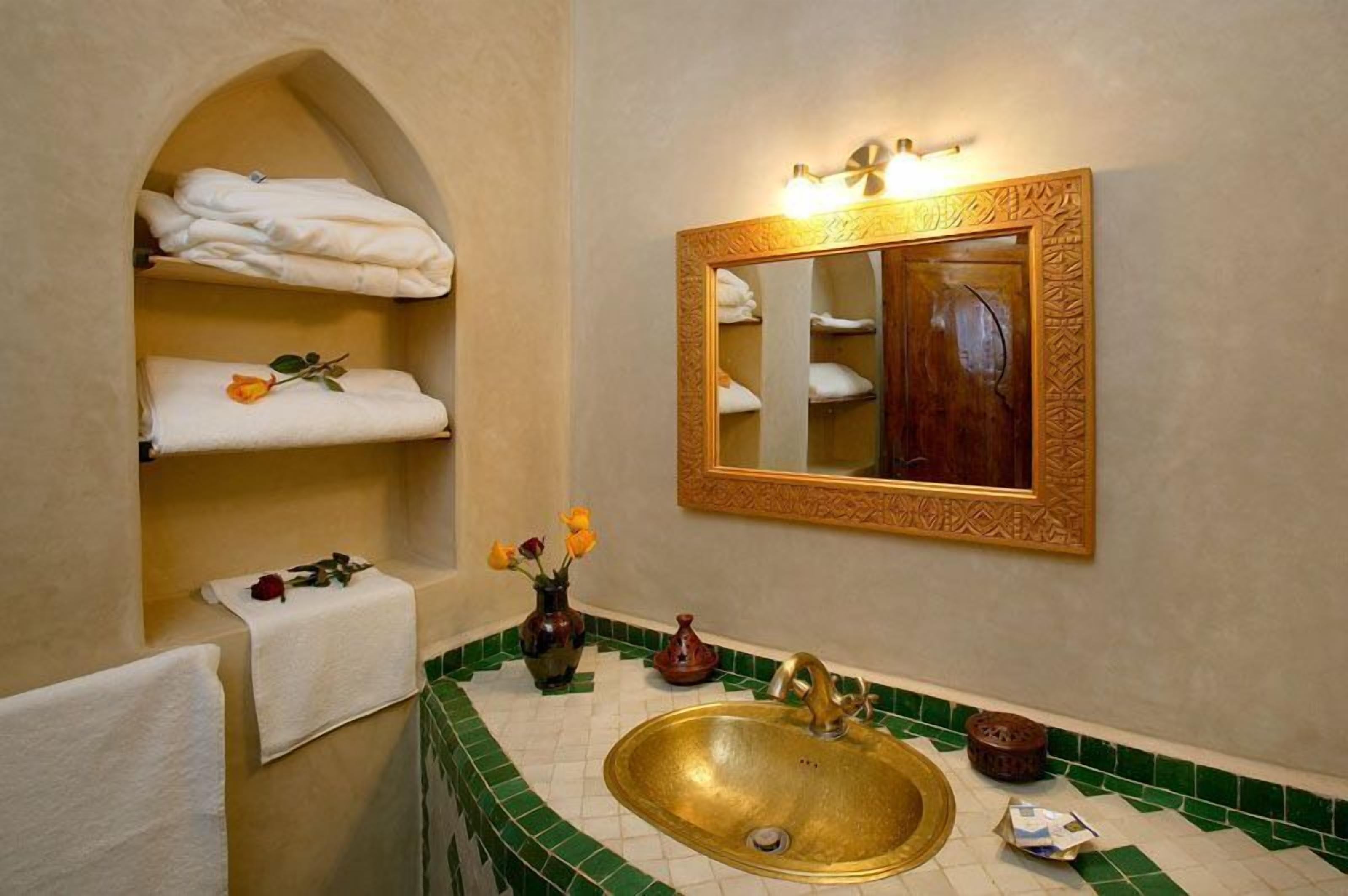 A bathroom with a green-tiled sink, a gold-framed mirror, and shelves with folded towels.