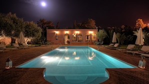 A rectangular swimming pool with a tiled deck, surrounded by lounge chairs and umbrellas, and a building with arched windows and warm lighting in the background. - Villa Dar Mya Palmiers (Marrakech)