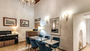 A dining area with a wooden floor, blue chairs, a table, a wooden cabinet, and wall-mounted artwork. - Little Queen Suite (Rome)