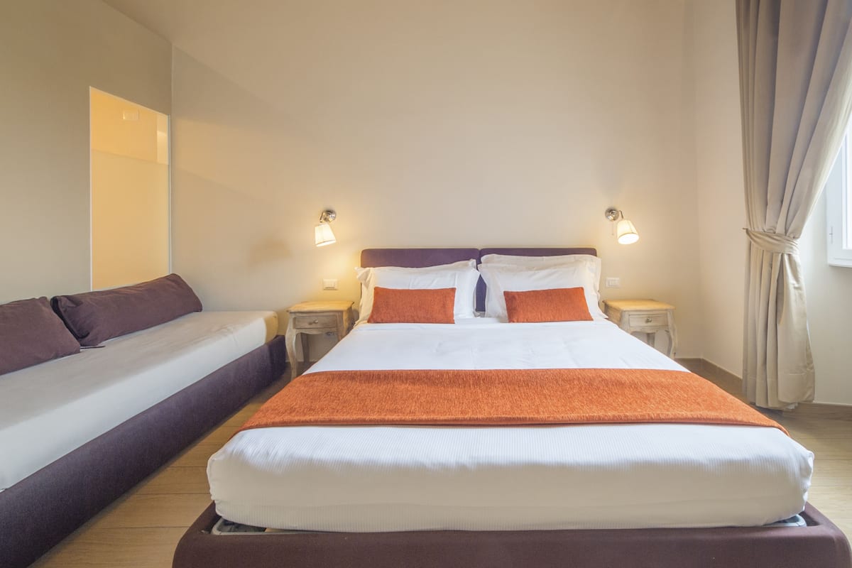 Ein Doppelbett mit orangefarbener Tagesdecke und zwei Kissen, mit Nachttischen mit Lampen an den Seiten, in einem Zimmer mit großem Spiegel und einem fenster mit Vorhängen.