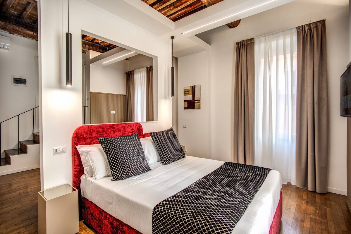 Ein modernes Schlafzimmer mit einem rot gepolsterten Kopfteil, einem großen Bett mit gemusterten Betttextilien, einem Flachbildfernseher und einer Holztreppe.