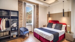 A modern bedroom with a red bed, a blue chair, a wardrobe, and a view of buildings through a window. - Little Queen Suite (Rome)