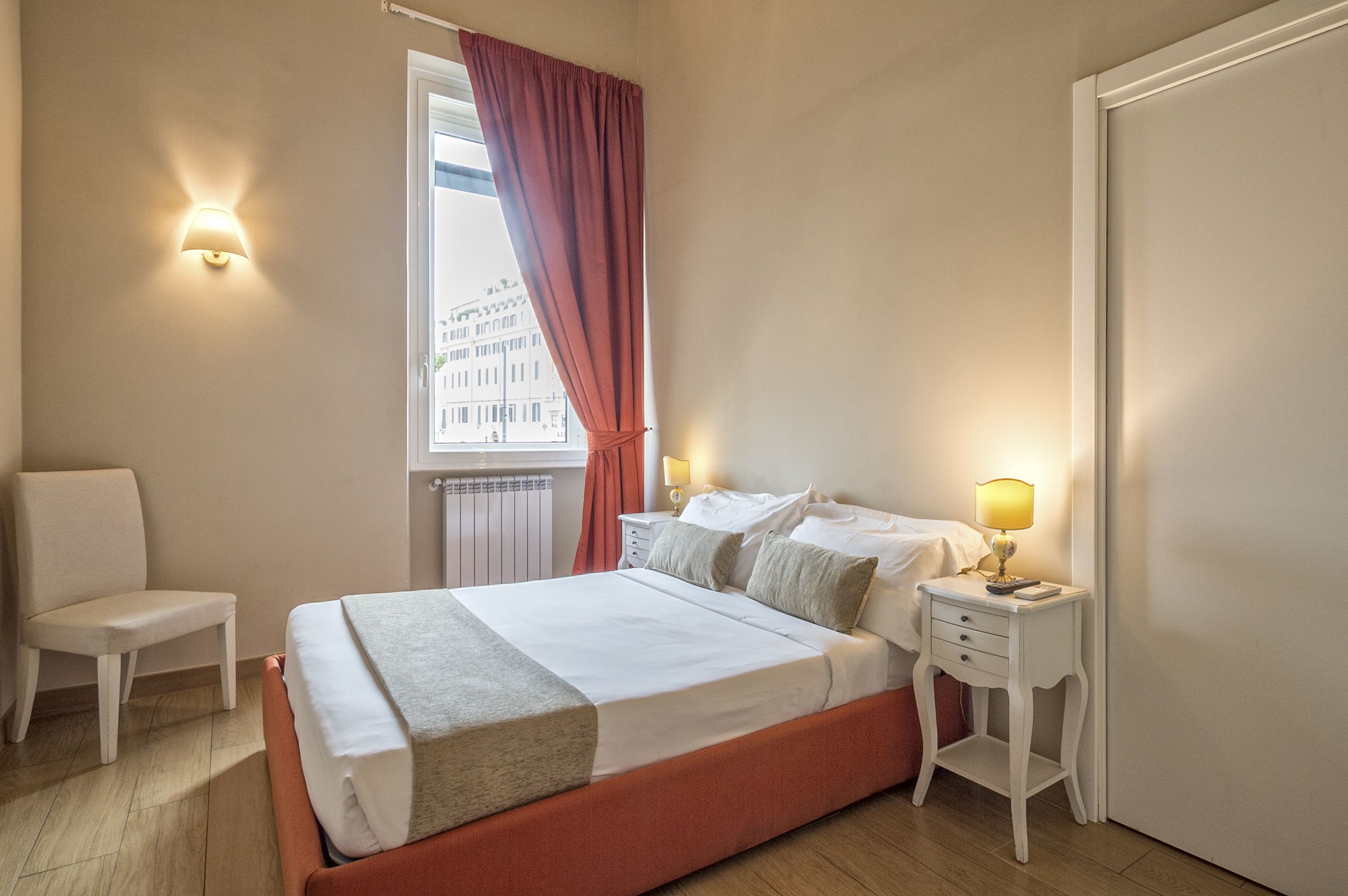 Um quarto com uma cama, uma cadeira, uma mesa de cabeceira com abajur, uma janela com cortinas e um radiador.