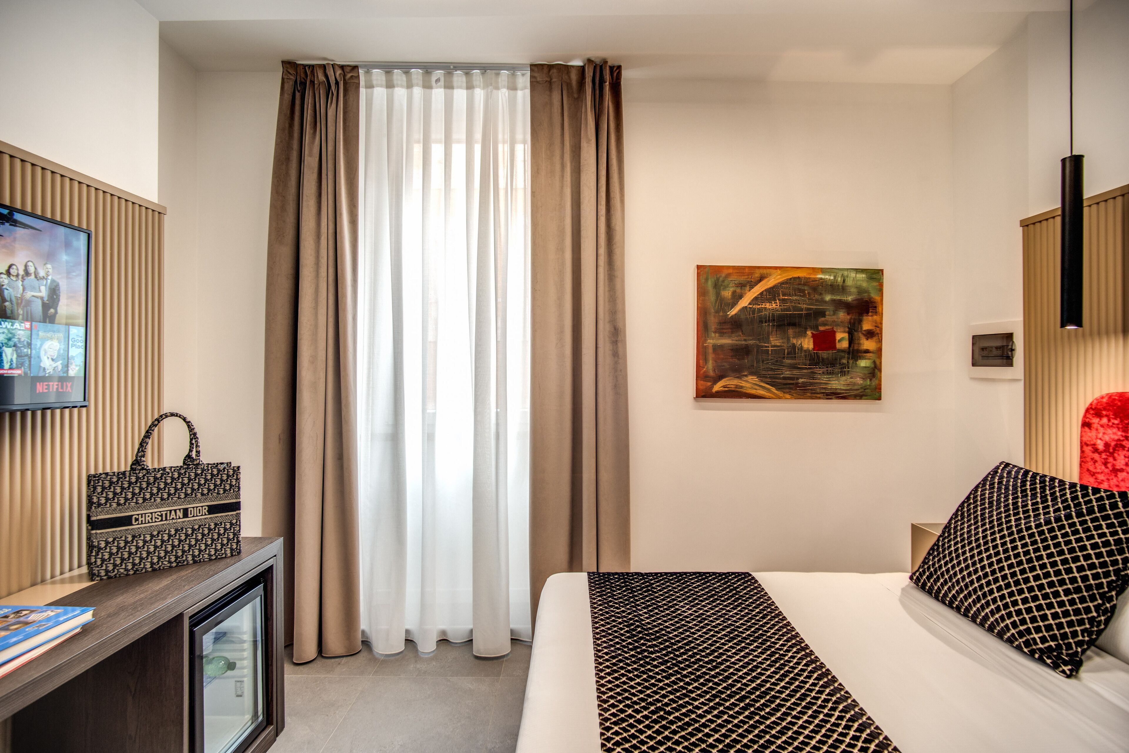 A hotel room with a bed, a television on a stand, a painting on the wall, and a window with curtains.