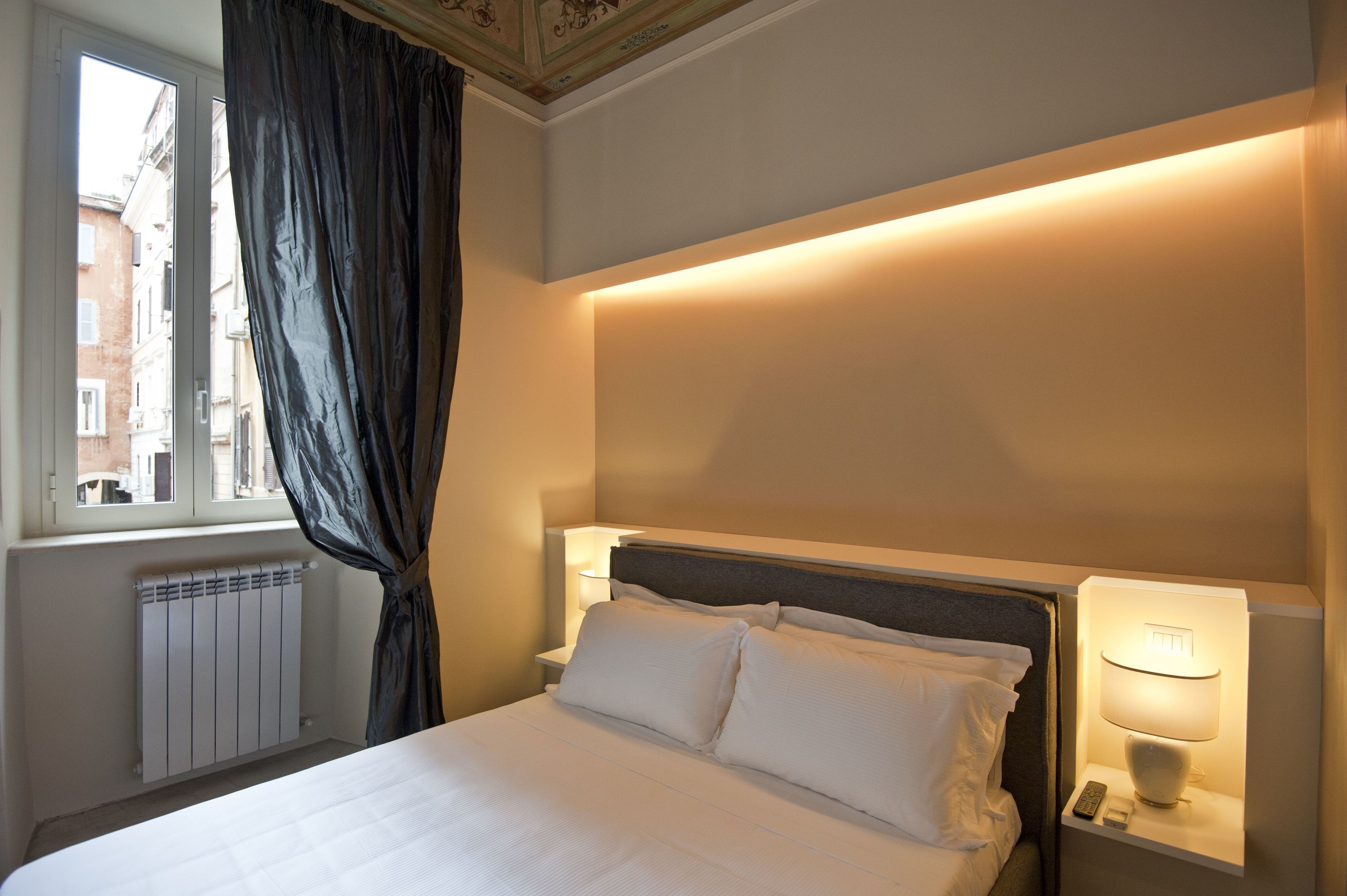 A hotel room with a bed, a nightstand, a lamp, a window with curtains, and a radiator.