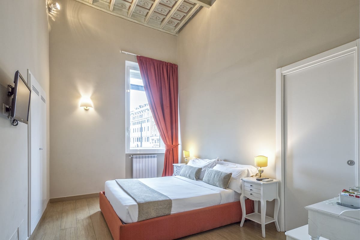 Ein Schlafzimmer mit Bett, Nachttischen, einem Fenster mit Vorhängen, einem Heizkörper und einer Deckenleuchte.