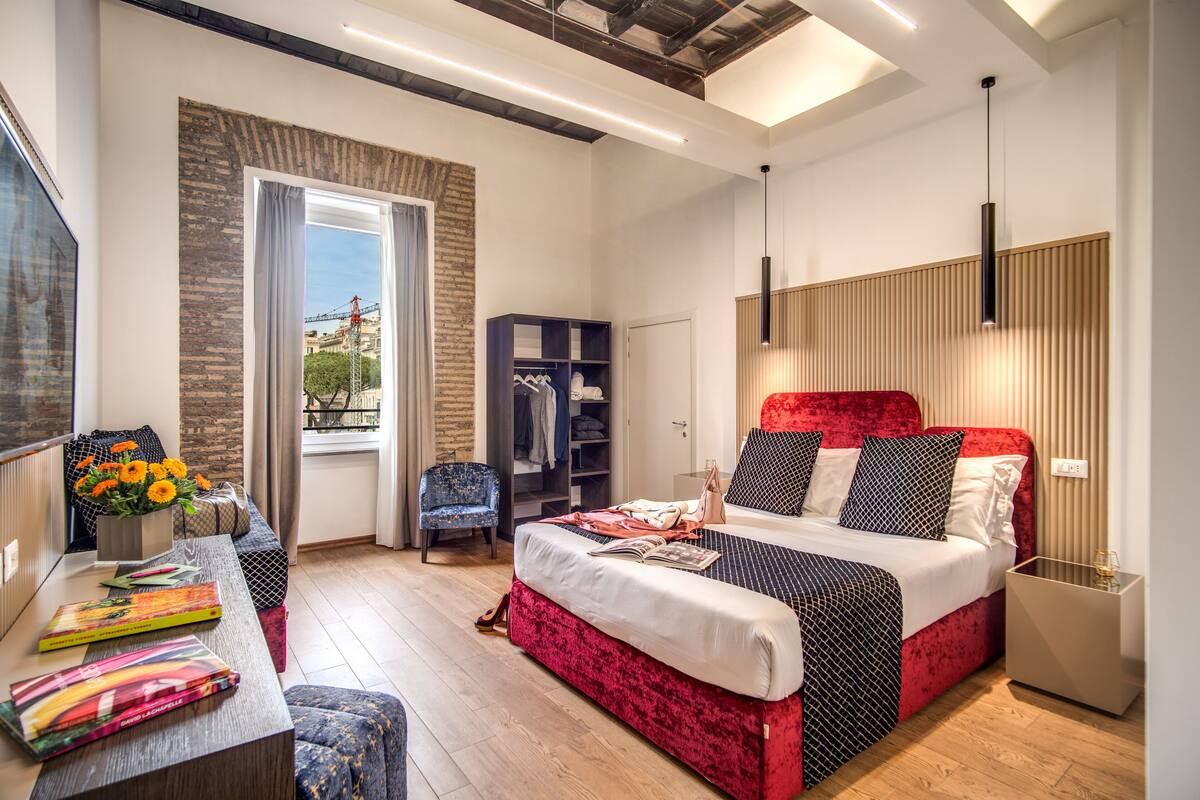 Ein modernes Schlafzimmer mit einem roten Bett, Holzboden, einem Bücherregal und einem Fenster mit Blick auf Gebäude.