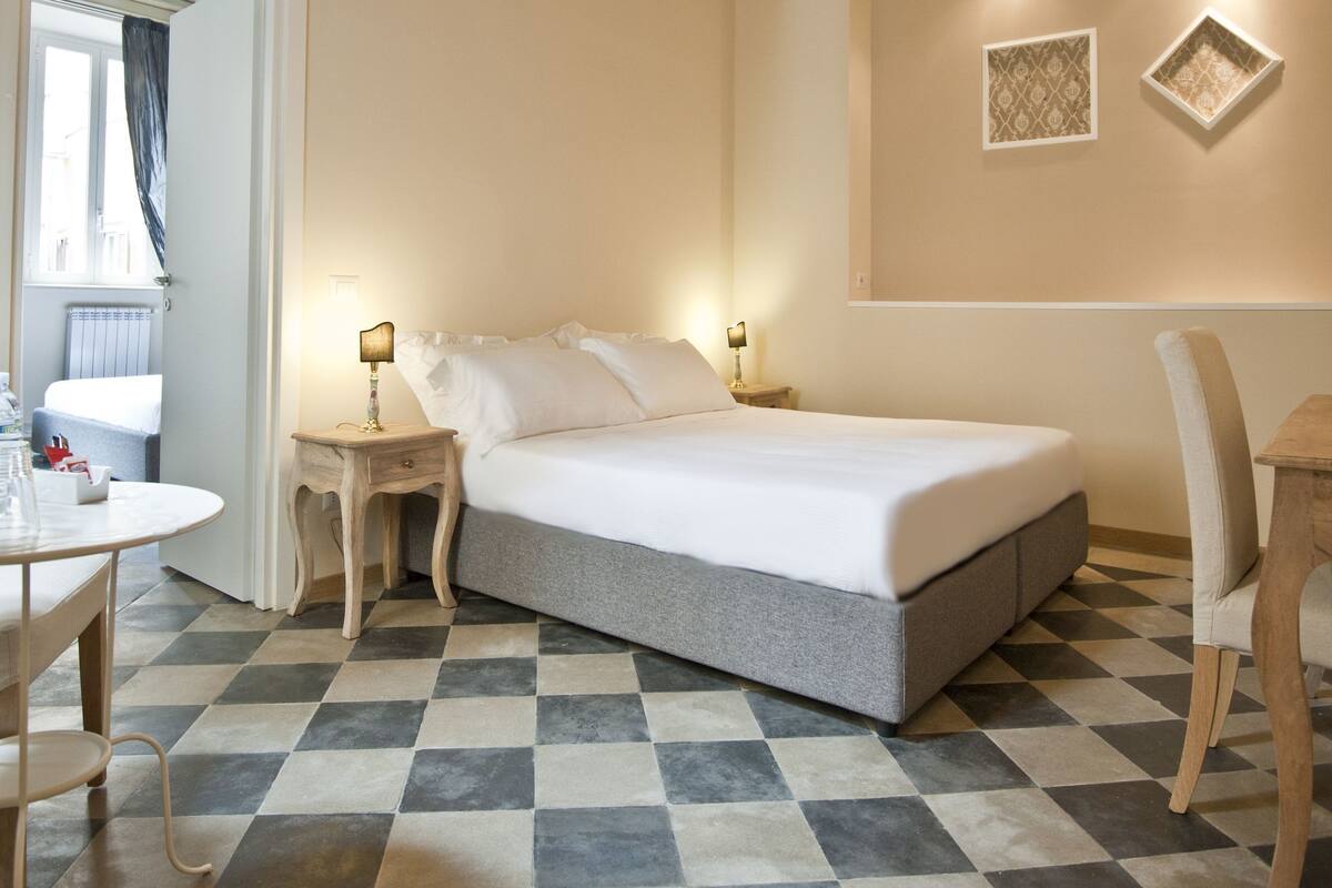 Ein Hotelzimmer mit Schachbrettmuster-Boden, einem Bett, einem Nachttisch mit Lampe, einem Sessel und einem kleinen Tisch mit Flasche und Gläsern.
