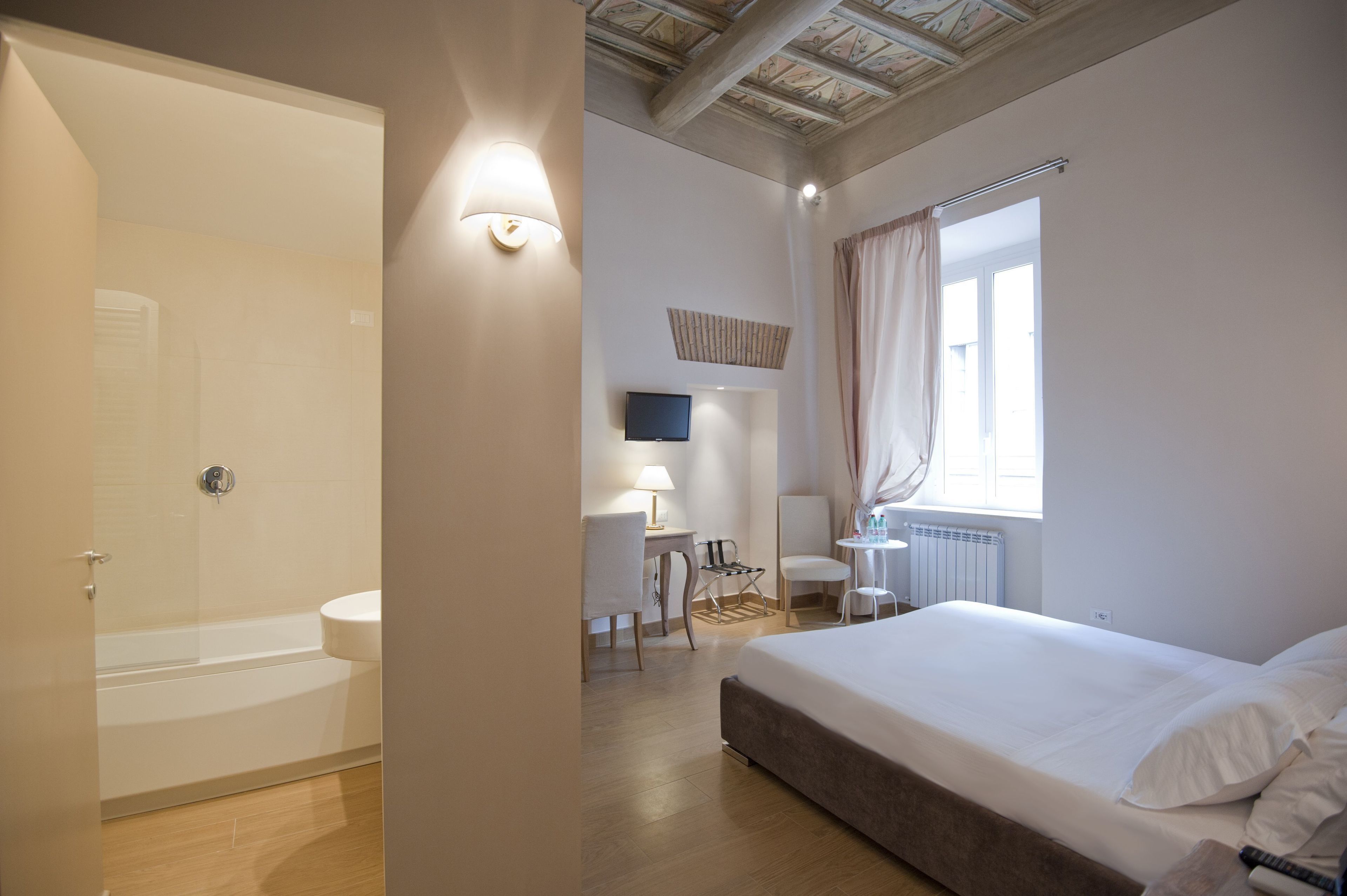 A hotel room with a bathtub, a desk with a chair, a small table with a lamp, and a bed with pillows.