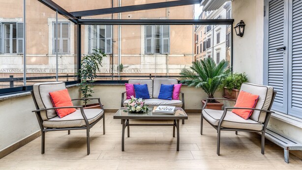 A balcony with two chairs and a table, a potted plant, and a view of a building with shutters. - Little Queen Suite (Rome)
