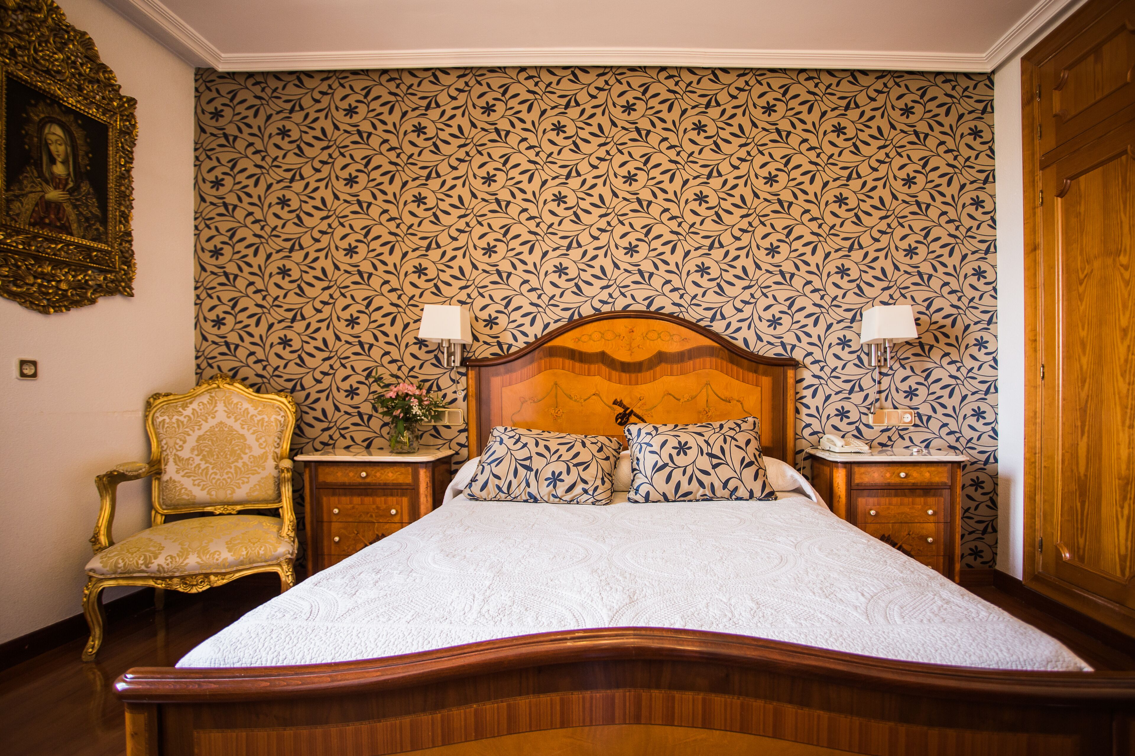 A bedroom with a large bed, a nightstand, a chair, and a wooden wardrobe.