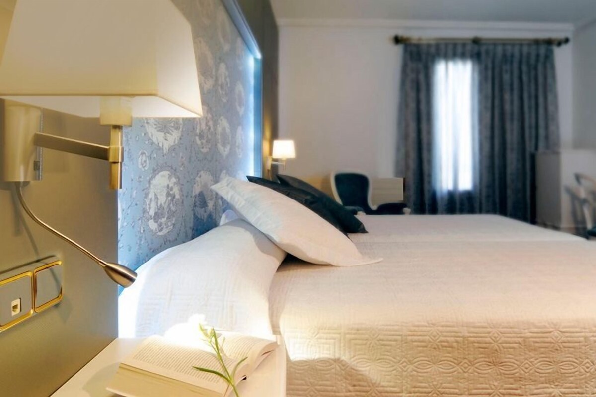 Habitación de hotel con una cama grande, un escritorio con silla, una lámpara y una ventana con cortinas.