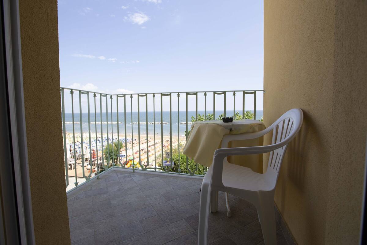 A balcony with a view of the beach, a table with a tablecloth, and a white plastic chair.