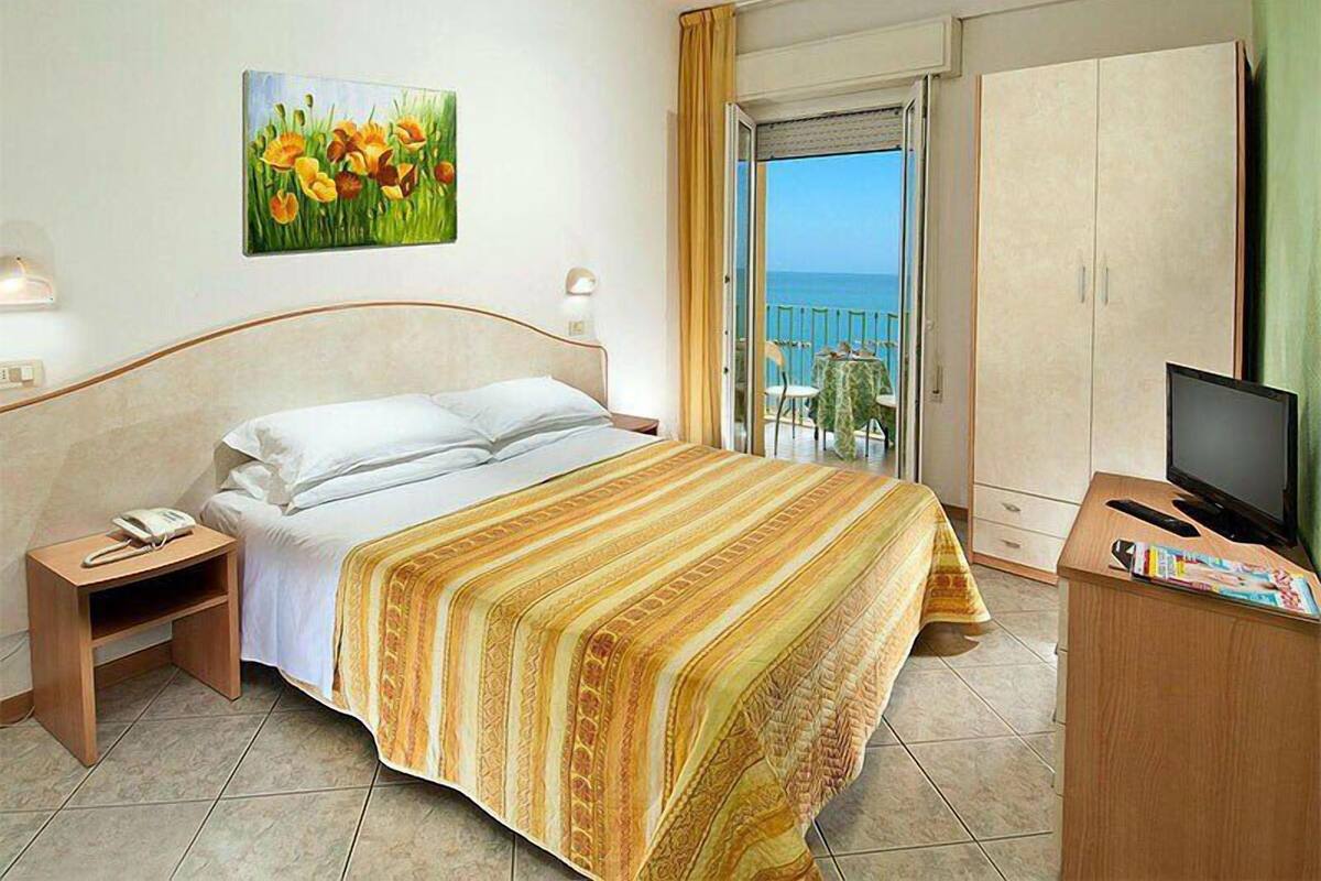 A hotel room with a bed, a TV, a desk, a chair, a table, a painting, and a balcony with a view.