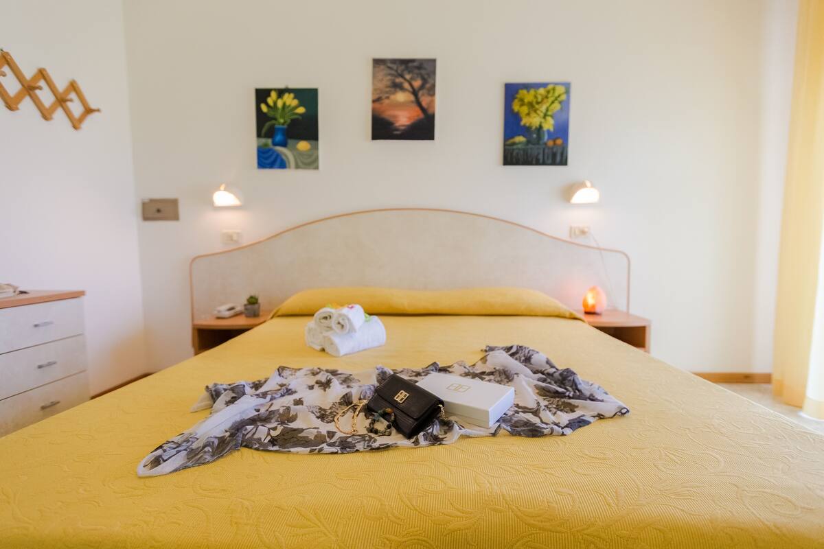 A neatly made bed with a yellow bedspread, a black handbag, and a white box on it. There are two bedside tables with lamps, a wall clock, and three framed paintings above the bed.
