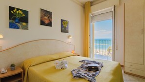 A hotel room with a bed, a nightstand, a lamp, a painting, a window with a view of the sea, and a door leading to a balcony. - Hotel Gloria (Gabicce Mare)