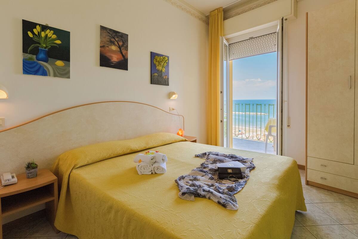 A hotel room with a bed, a nightstand, a lamp, a painting, a window with a view of the sea, and a door leading to a balcony.