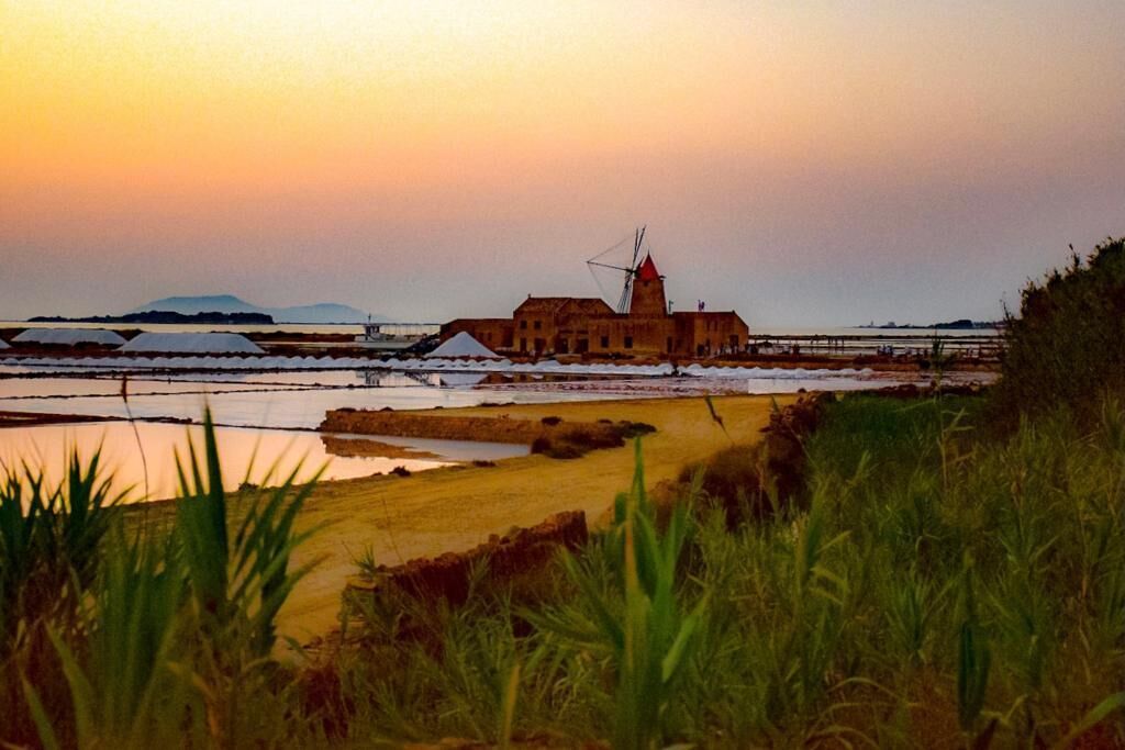 A coastal building with a windmill, surrounded by saltpans and water bodies, during sunset.