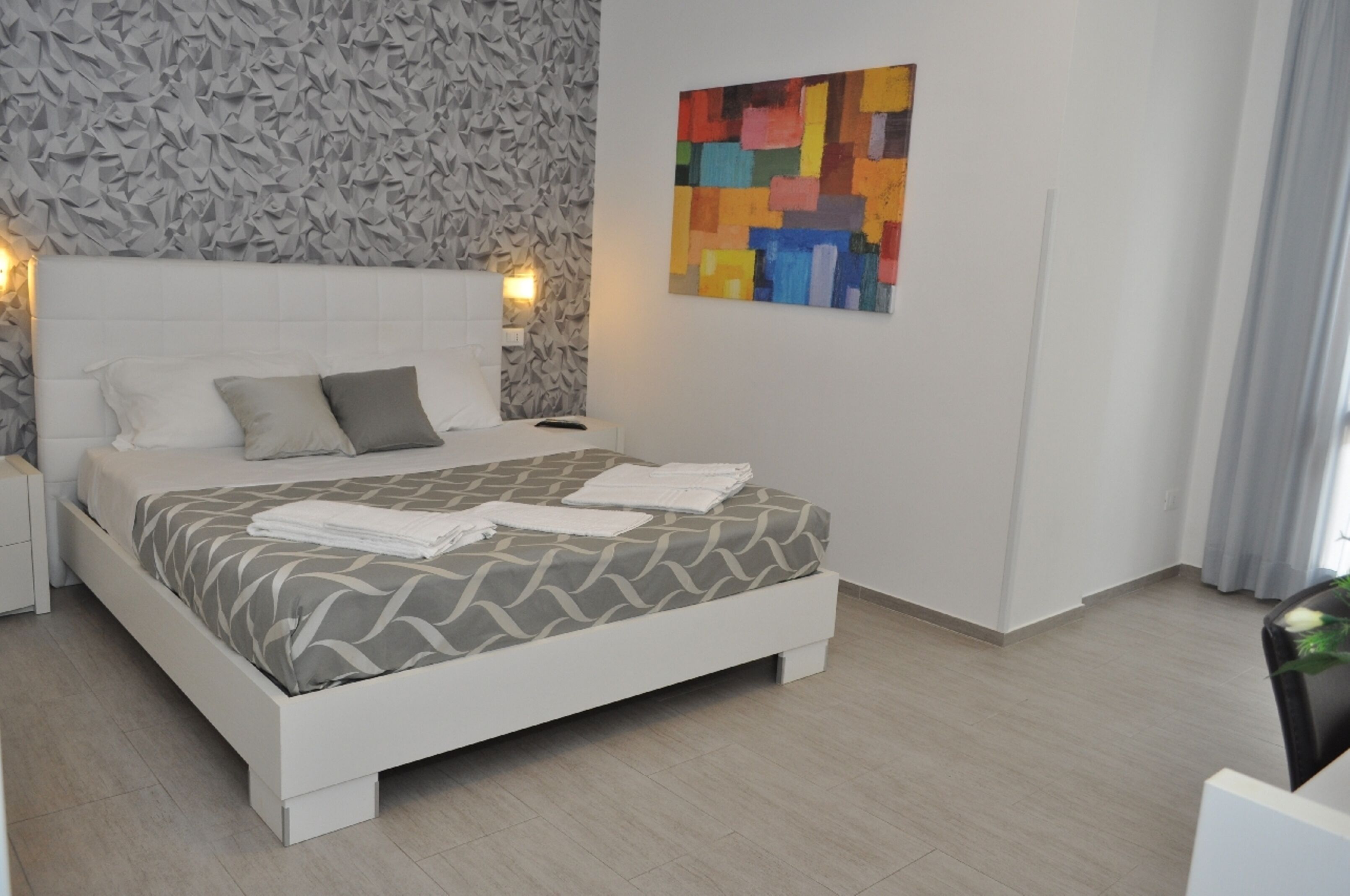 A modern bedroom with a white bed, grey patterned bedding, a colorful abstract painting, and a textured wall.
