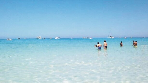 Een strand met helder turquoise water, verschillende boten en mensen die zwemmen.