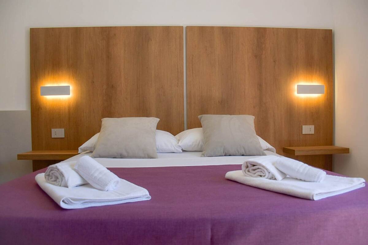 Un lit double avec une couvre-lit violette, deux oreillers blancs et deux serviettes blanches pliées sur le lit. Il y a deux tables de chevet avec des appliques murales.