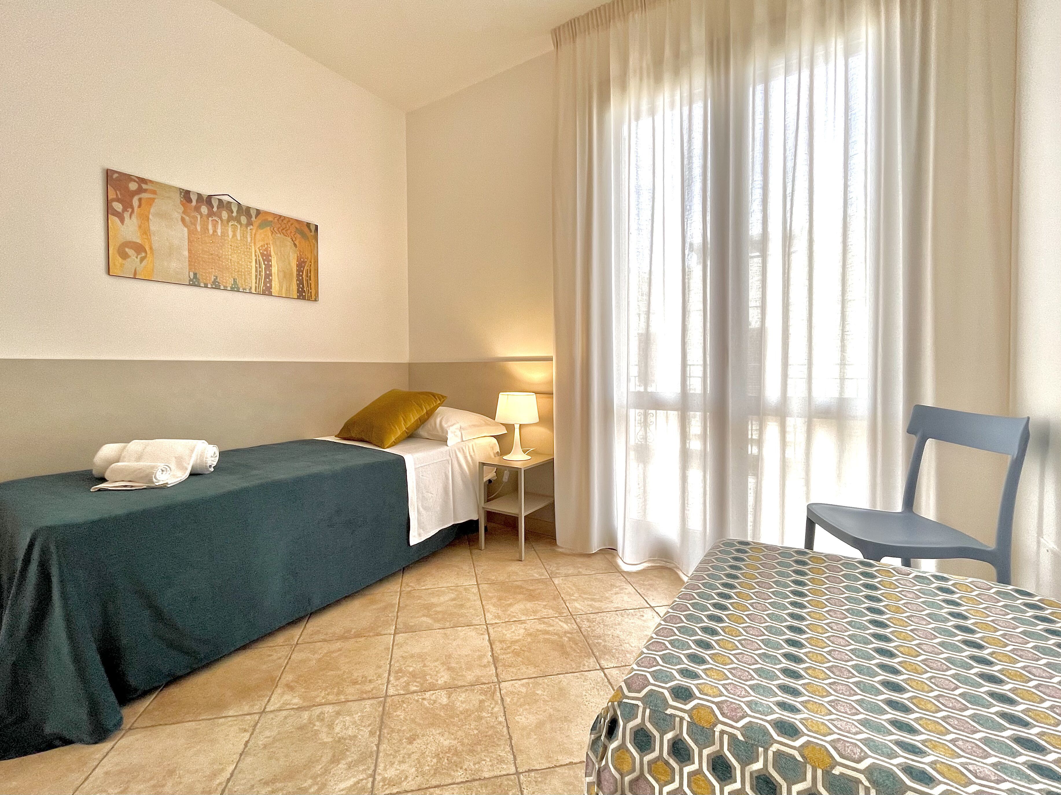 A hotel room with two beds, a nightstand, a lamp, a chair, and a painting on the wall.