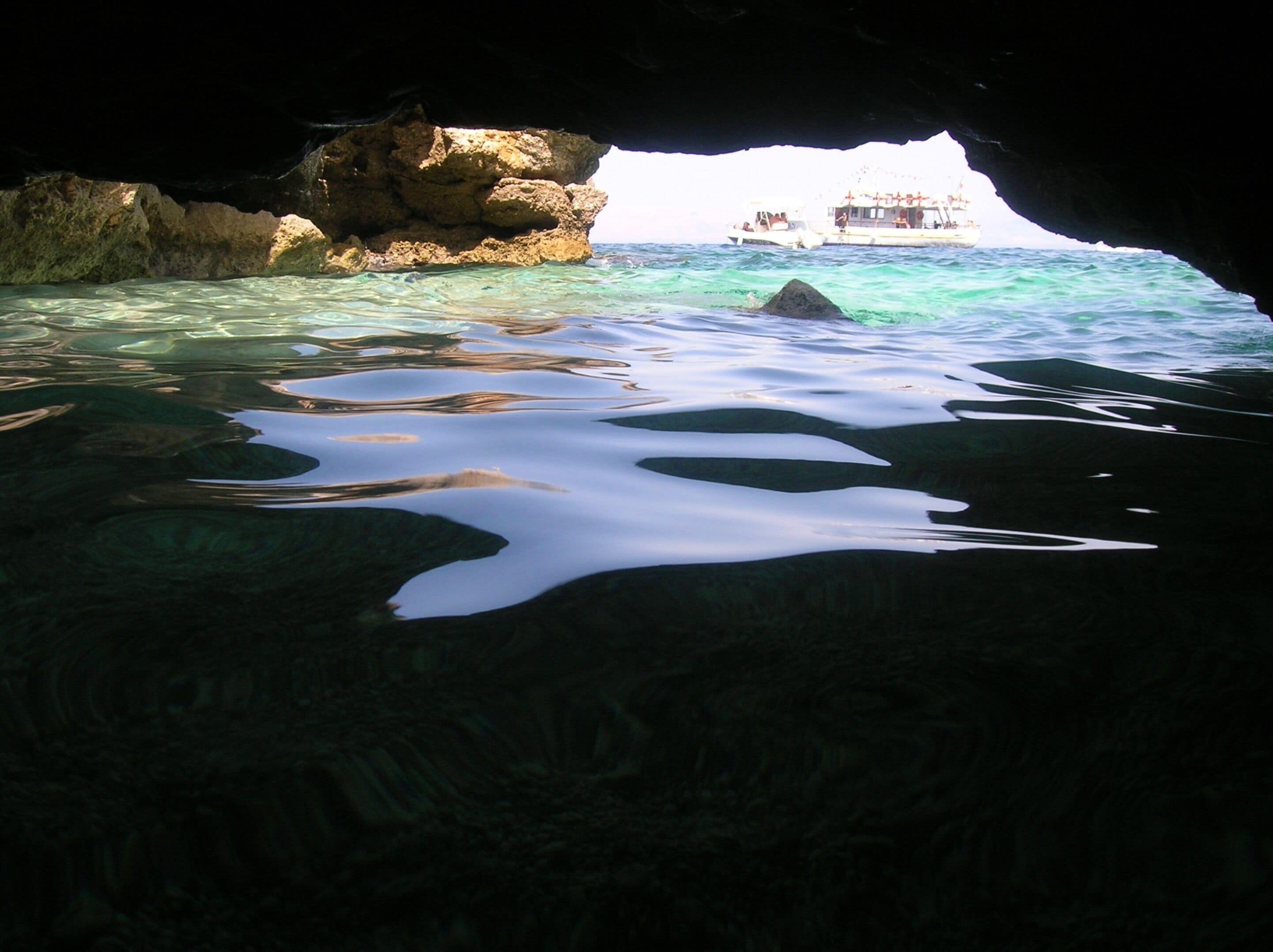 A cave entrance with clear water leading to a boat and a building in the distance.
