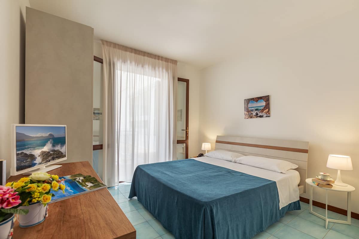 Une chambre d’hôtel avec un lit, des lampes de chevet, un bureau avec un écran d’ordinateur et un vase de fleurs, ainsi qu’une fenêtre avec des rideaux.