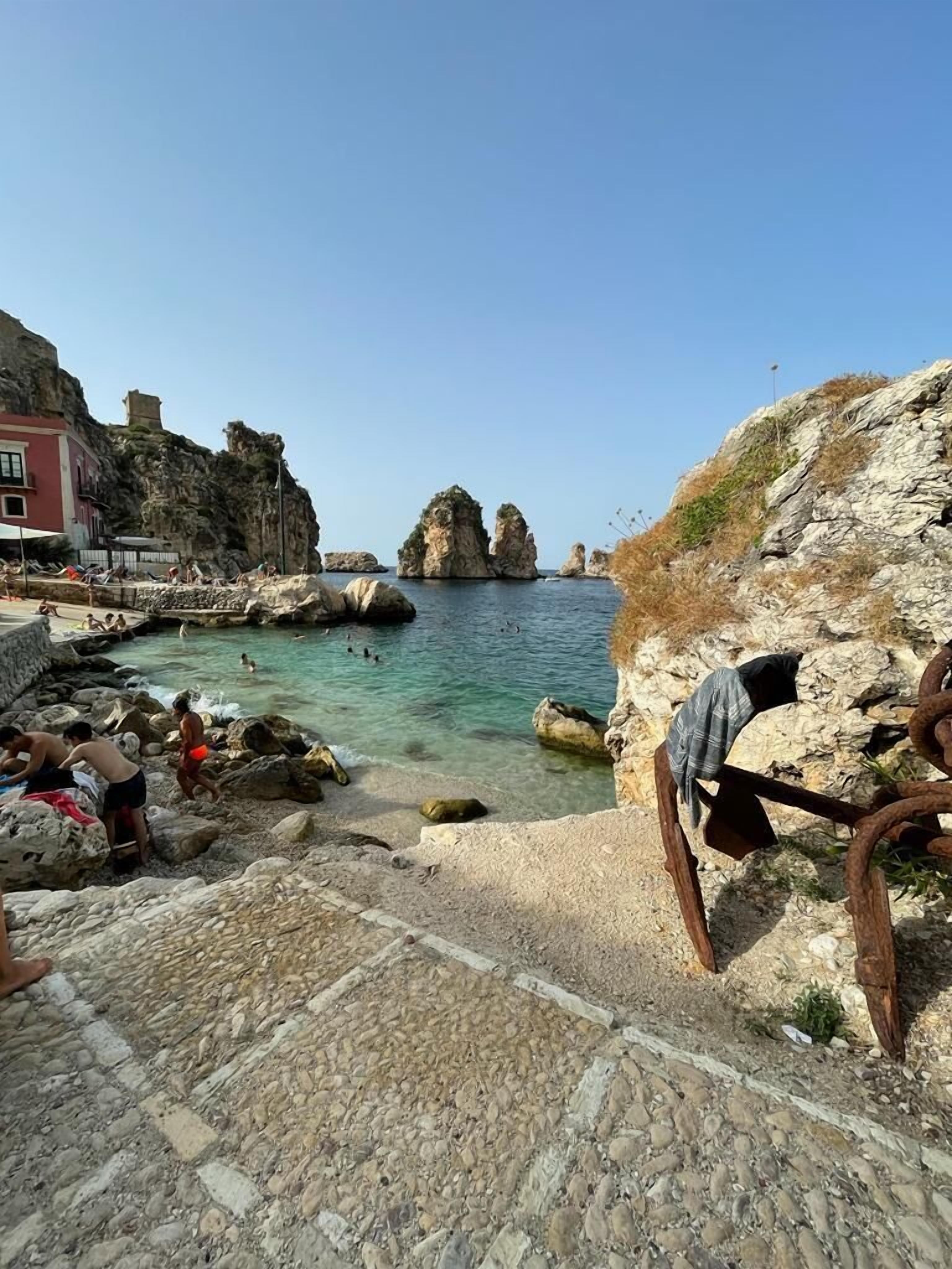 A rocky coastal area with clear water, a paved walkway, and a building with a red facade.