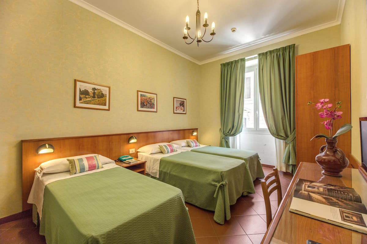 A hotel room with two beds, a wooden headboard, a dining table with a vase of flowers, and a window with curtains.