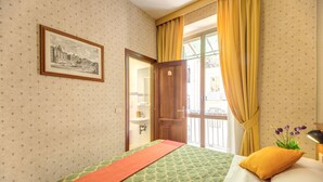 A hotel room with a bed, a framed picture on the wall, a window with yellow curtains, and a view of the outside. - Hotel Corona (Rome)