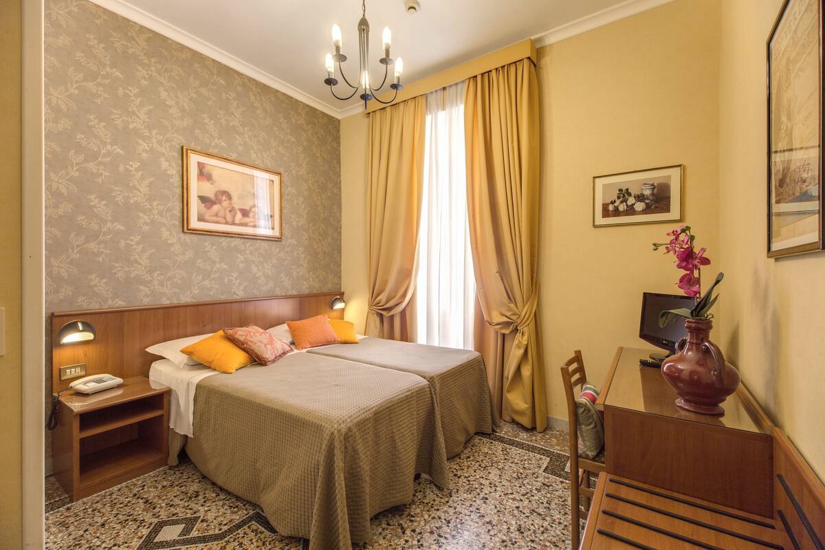 A hotel room with two beds, a desk, a chair, a telephone, and a vase with flowers.