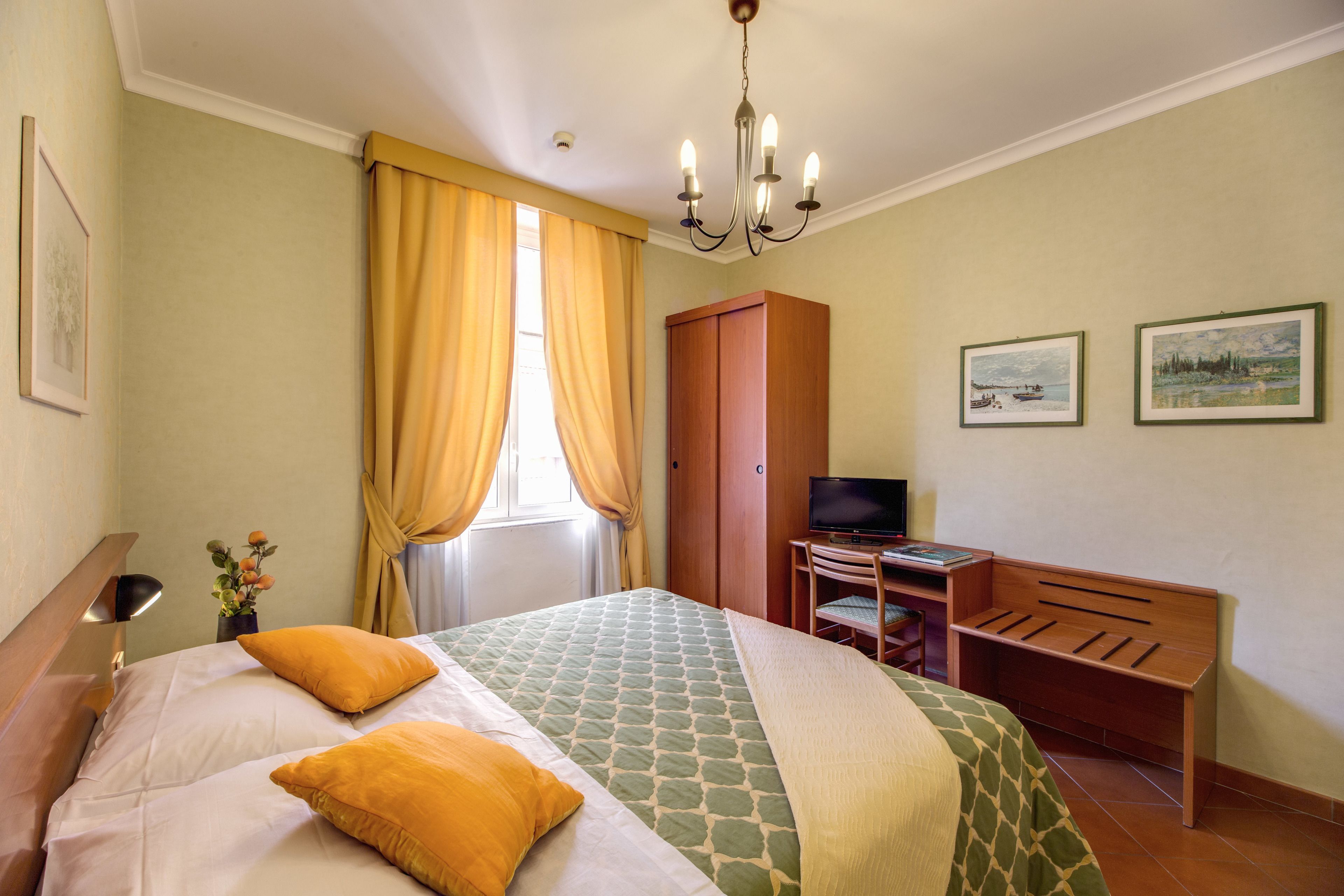Quarto de hotel com cama, escrivaninha com computador, televisão e duas quadros na parede.