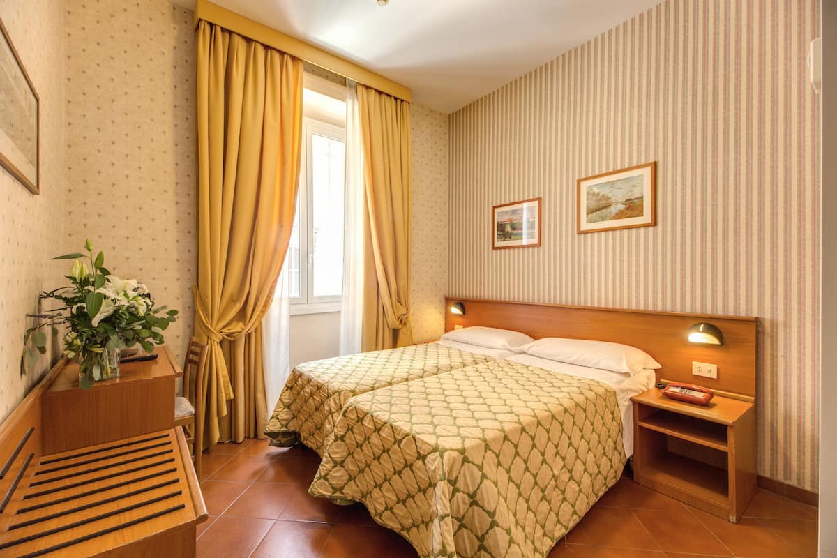 A hotel room with a bed, a nightstand, a lamp, a window with curtains, a plant, and a framed picture on the wall.