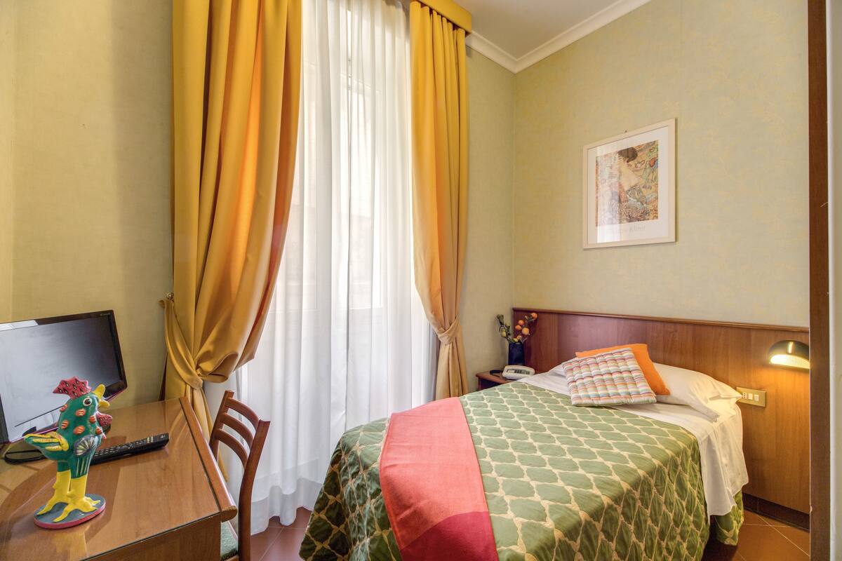 A hotel room with a bed, a desk with a computer, a chair, a lamp, and a painting on the wall.