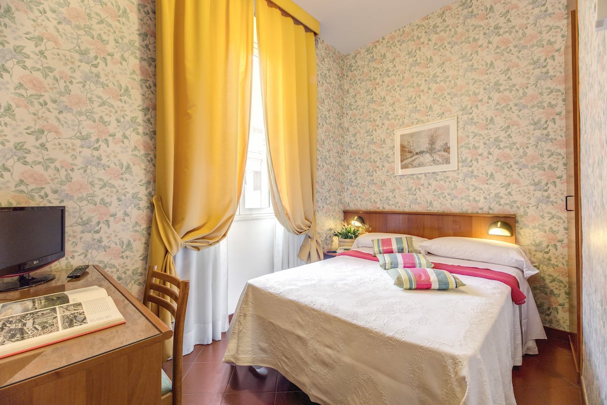 A hotel room with a bed, a desk with a computer, and a window with curtains.