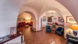 A spacious living area with a high arched ceiling, a red sofa, a blue armchair, and a small table with a lamp. - Hotel Corona (Rome)