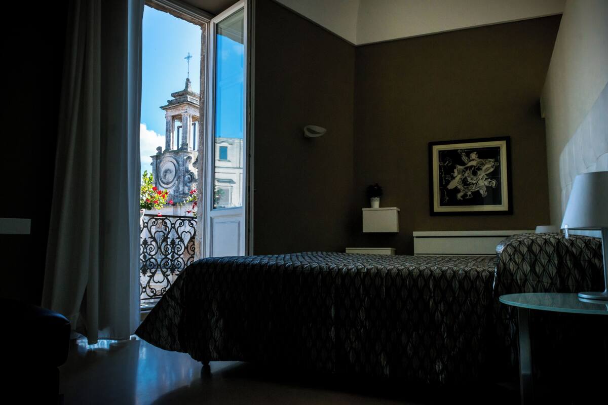 Una habitación de hotel con cama, una mesita, una mesita de noche, un cuadro en la pared y vistas a un edificio a través de una ventana.
