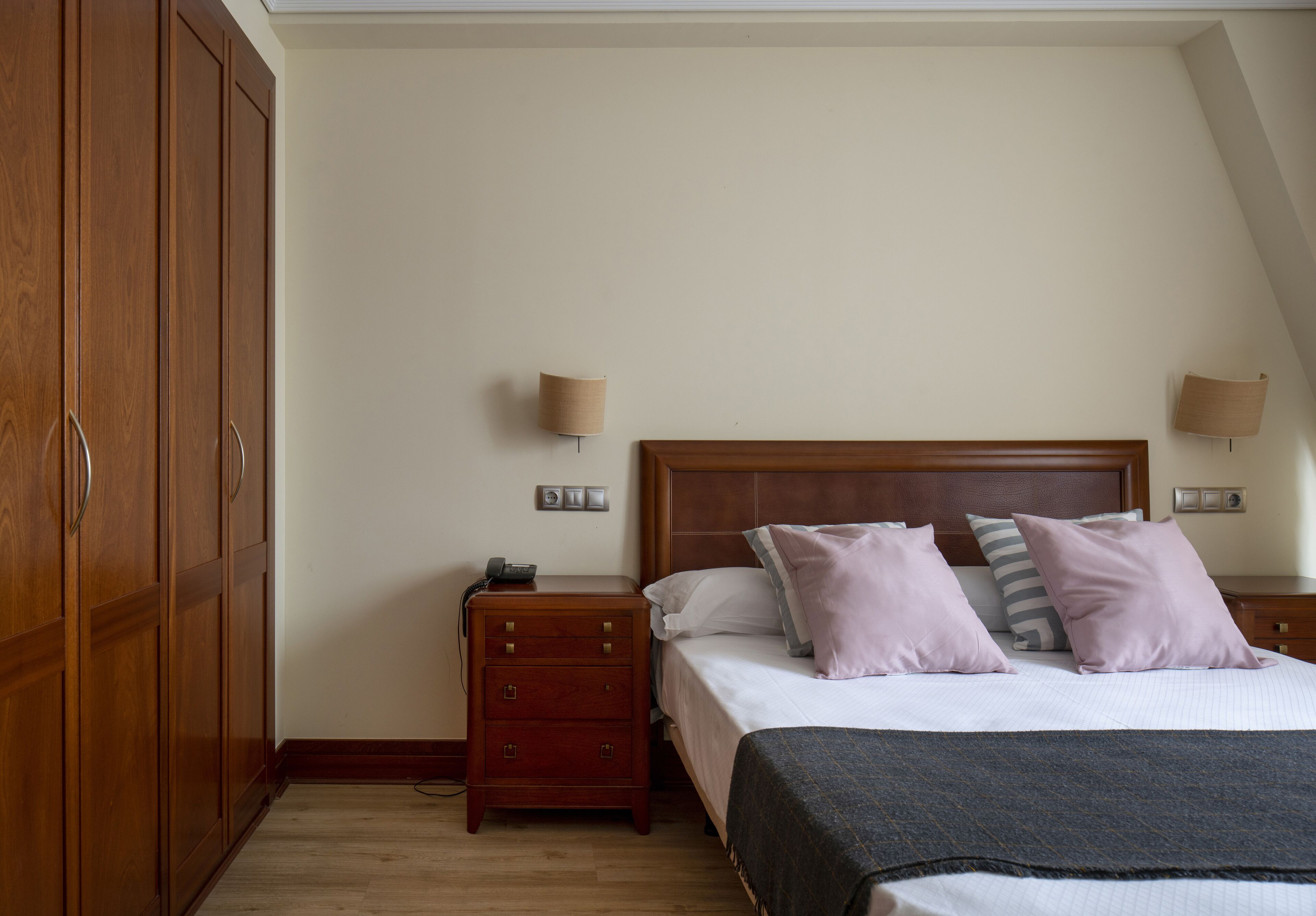 Un dormitorio con un armario de madera, una mesita de noche con lámpara, una cama con almohadas y un atril.