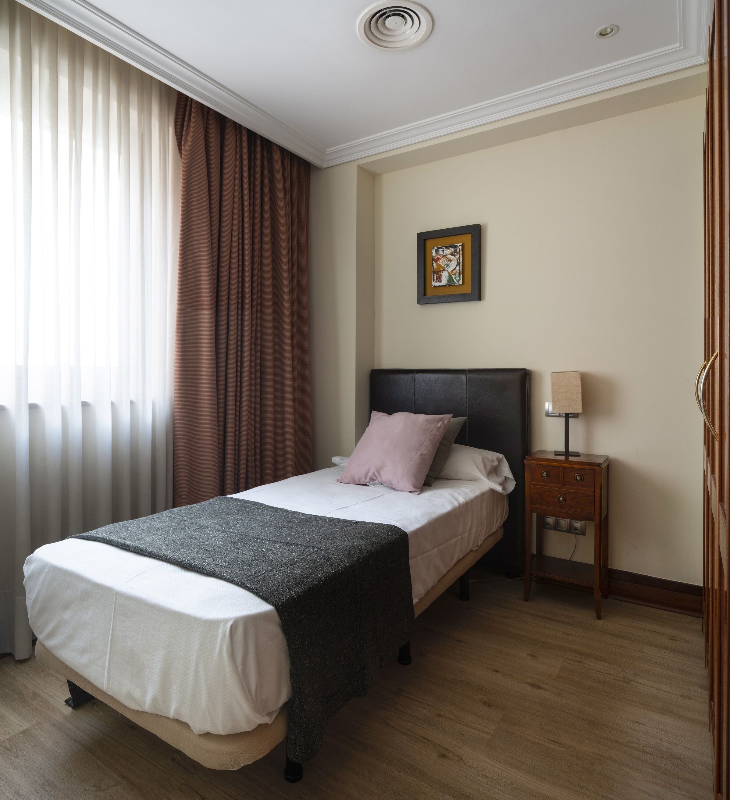 Una habitación de hotel con una cama, una mesita de noche, una lámpara, un cuadro en la pared y una ventana con cortinas.