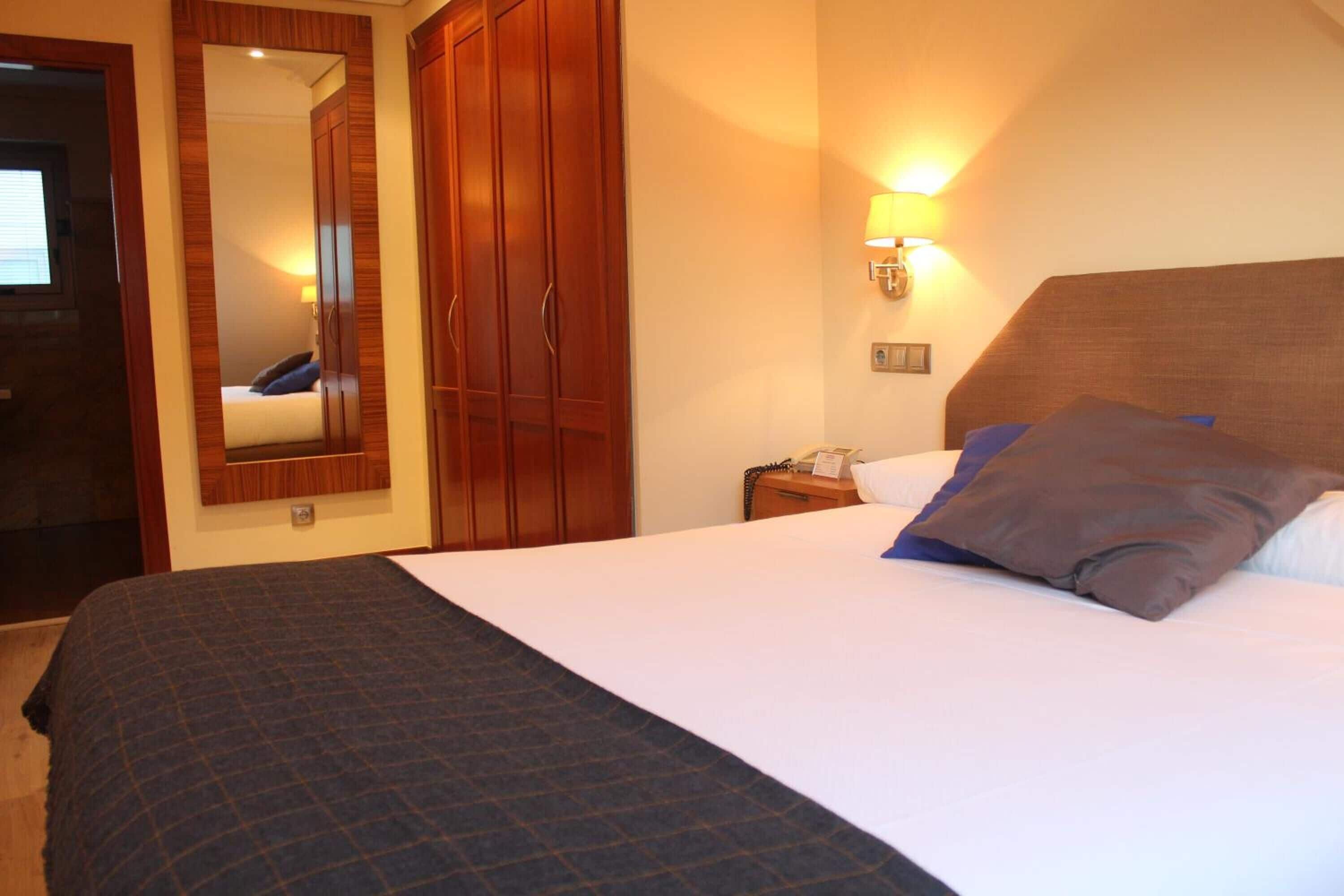 A hotel room with a large bed, wooden wardrobe, a mirror, and a wall-mounted lamp.