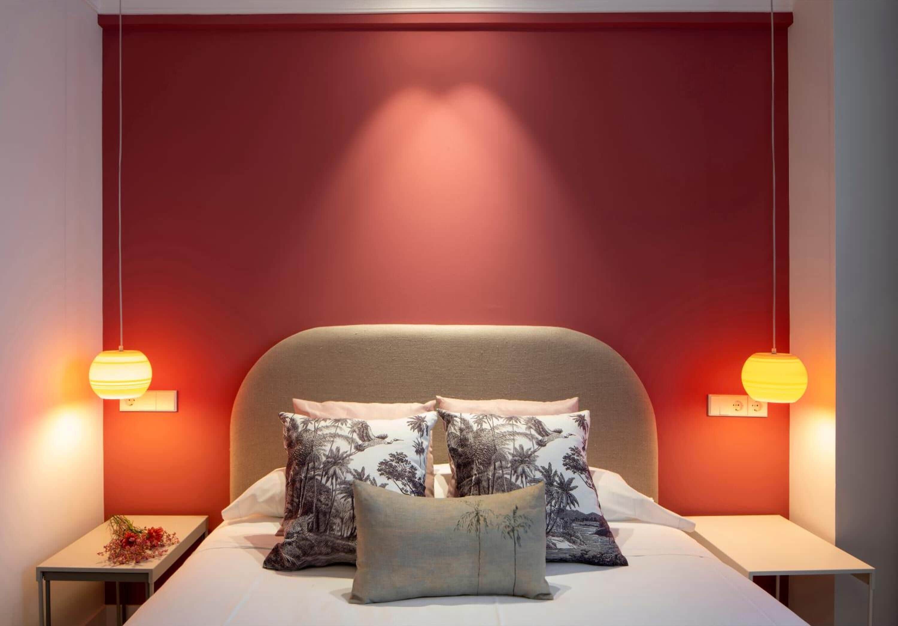 A bedroom with a red wall, a beige headboard, and two bedside lamps. There are two pillows with a tropical print and a single plain pillow.