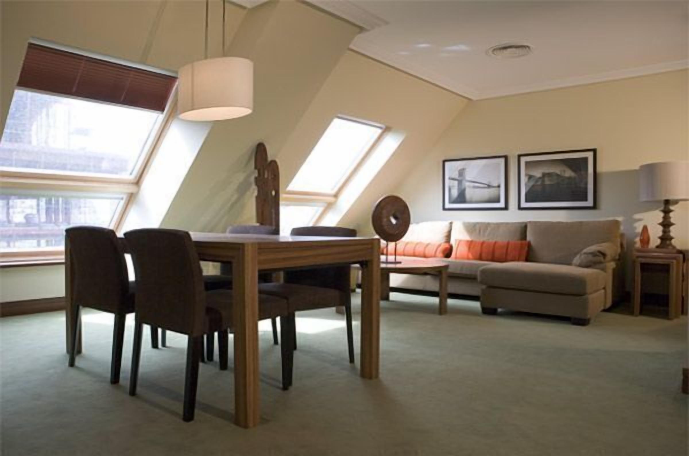 A modern living room with a dining table, chairs, a sofa, and a coffee table. There are two windows with blinds, a ceiling fan, and framed pictures on the wall.