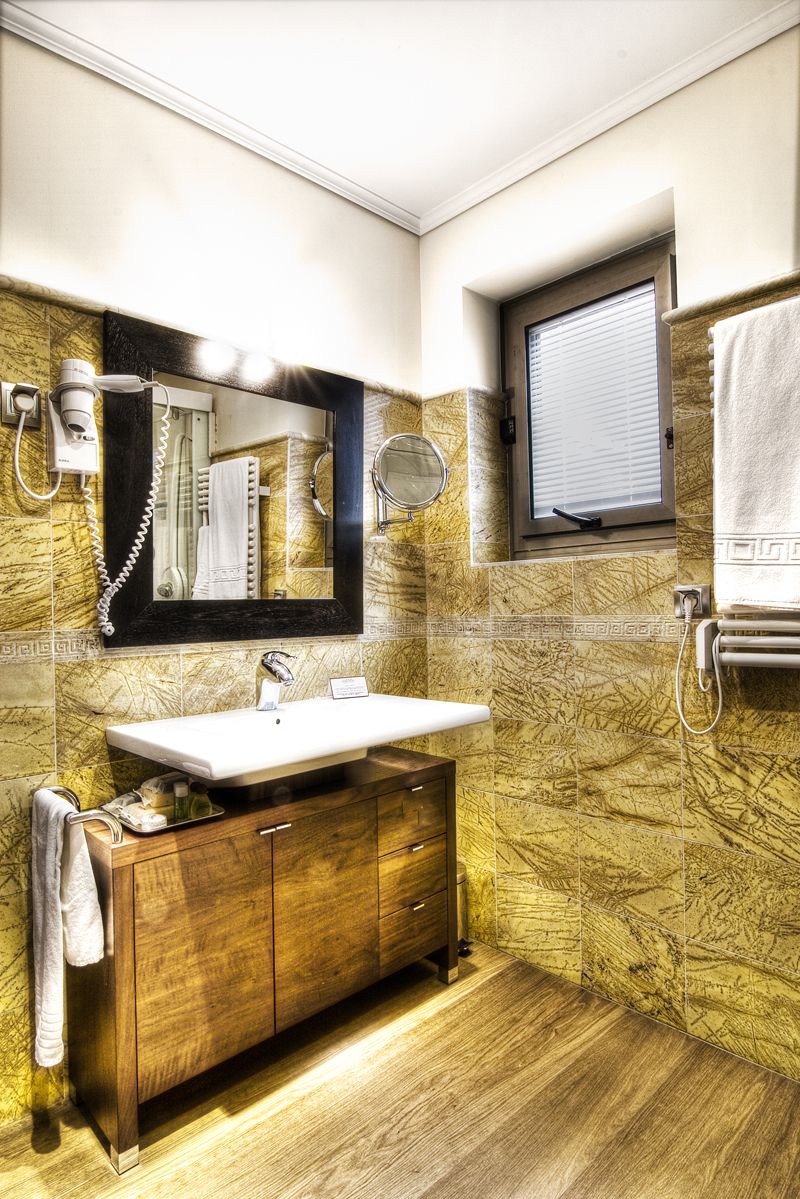 A bathroom with a marble wall, a wooden vanity, a sink, a mirror, a towel rack, and a window with blinds.