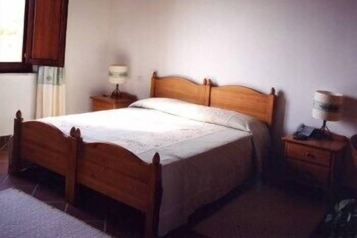 Una cama de madera con colcha blanca en una habitación con ventana, una mesita de noche con lámpara y una pequeña mesa con lámpara.