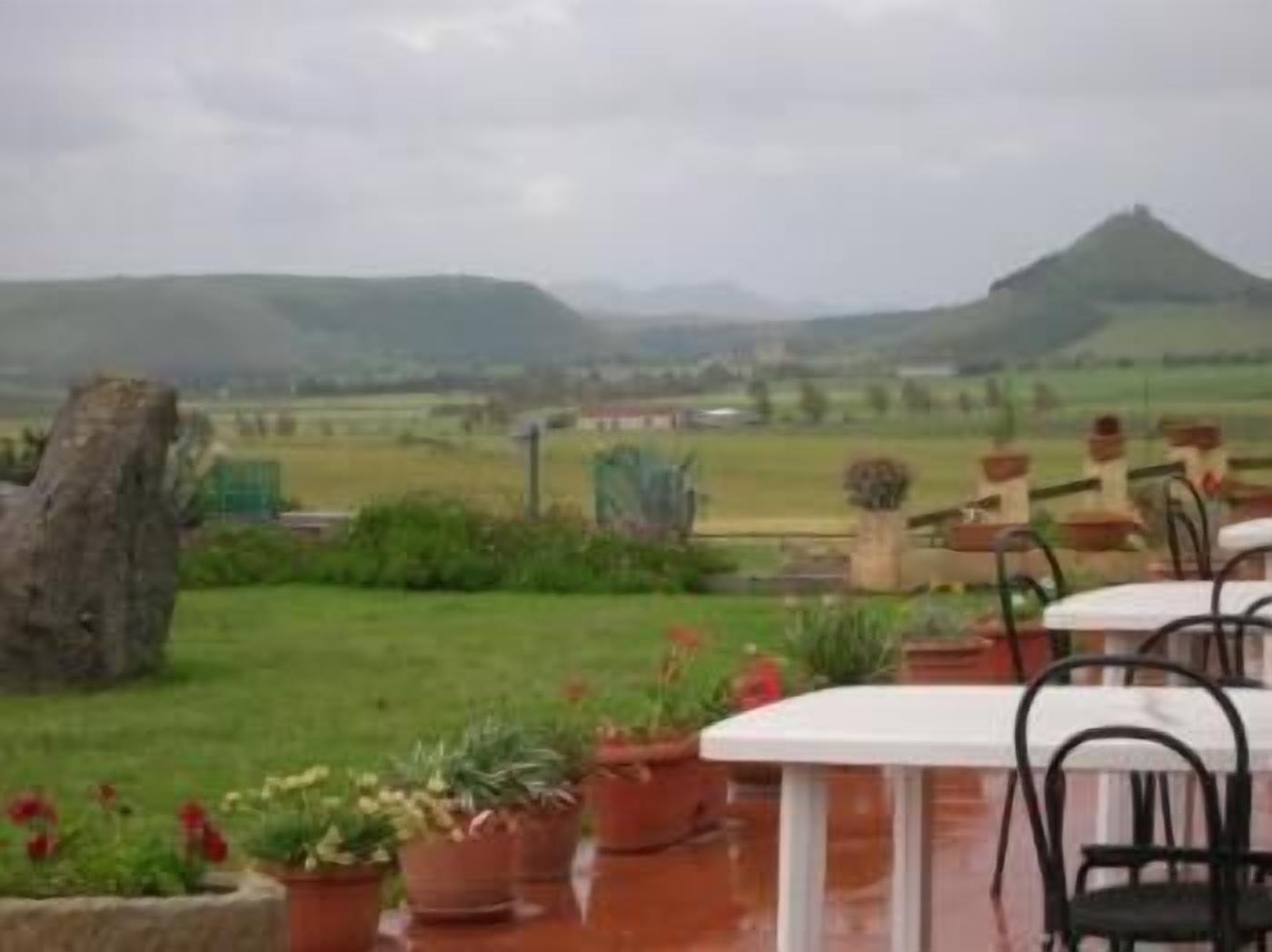 An outdoor seating area with tables and chairs, surrounded by potted plants and a view of distant hills.