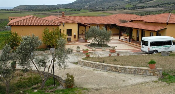 A yellow building with a red-tiled roof, a white van parked in front, and a stone wall surrounding the property. - Hotel Su Nuraxi (Barumini)