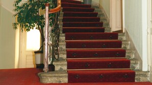 A grand staircase with red carpeted steps, a wooden handrail, and a potted plant at the top. - Hotel Heluan (Karlovy Vary)