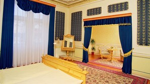 A room with a large bed, a wooden nightstand, a mirror, a chair, a small table, and a patterned rug. - Hotel Heluan (Karlovy Vary)