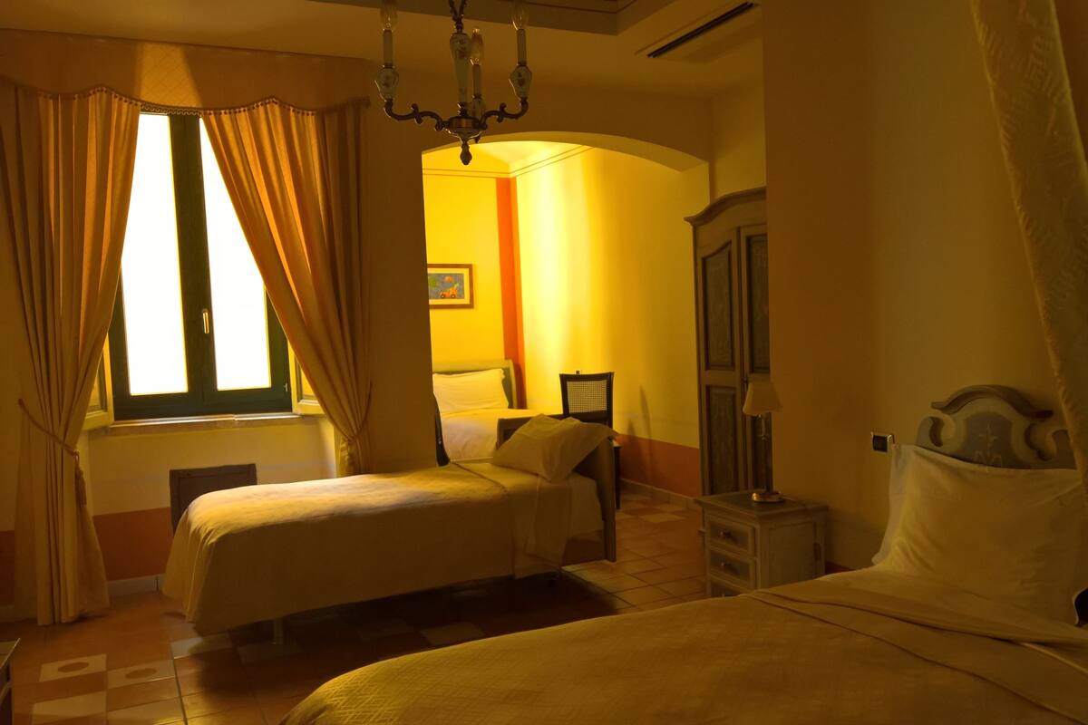 Une chambre d’hôtel avec deux lits, un lustre, une fenêtre avec des rideaux et une porte dotée d’un miroir.